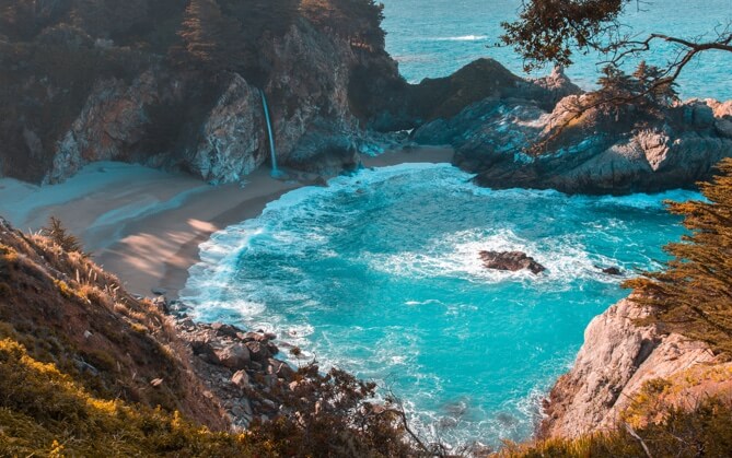 A small beach surrounded by cliffs, ocean, and a small waterfall.